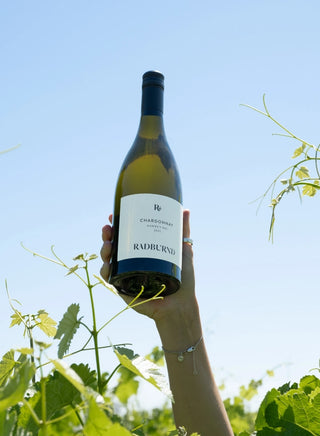 A bottle of Radburnd Cellars Hawke's Bay Chardonnay 2021 vintage raised against a summer sky in the vineyard.